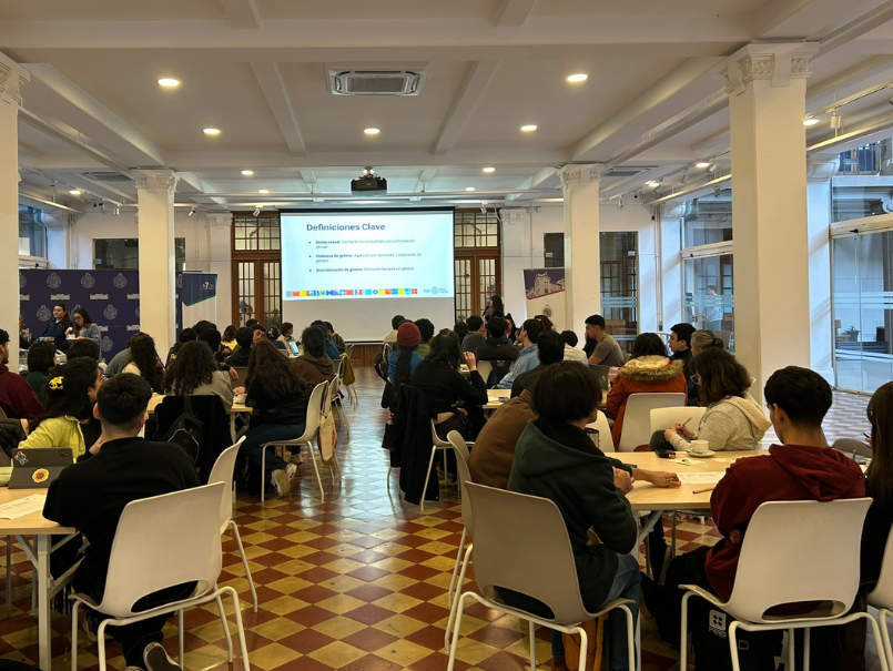 Asistentes de jornada están sentados en grupos, distribuidos en Salón Centenario de la PUCV. 